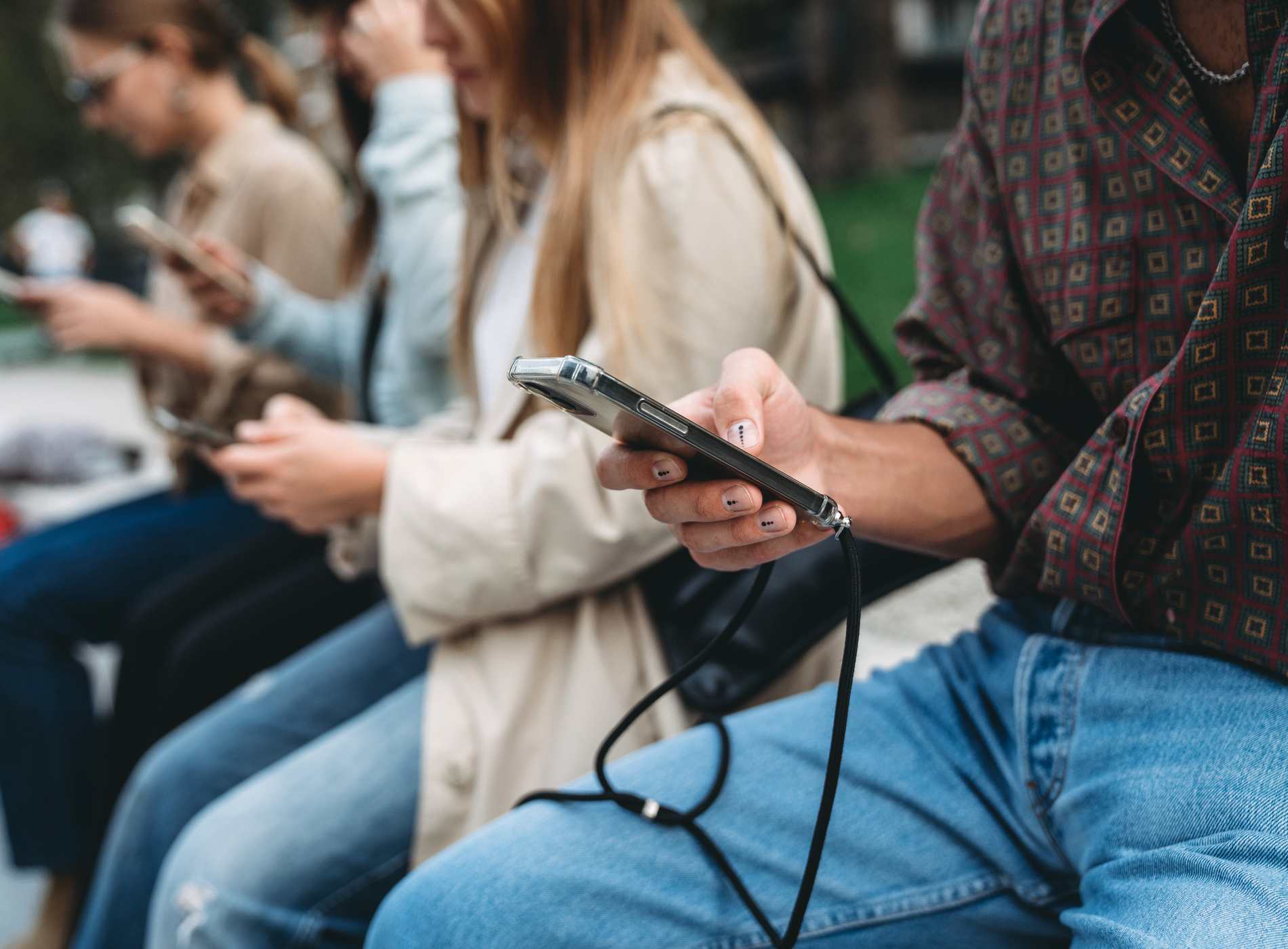 Four friends are using smart phones while they are sitting in a public park in the city. Detail of the hands holding the mobile phones.