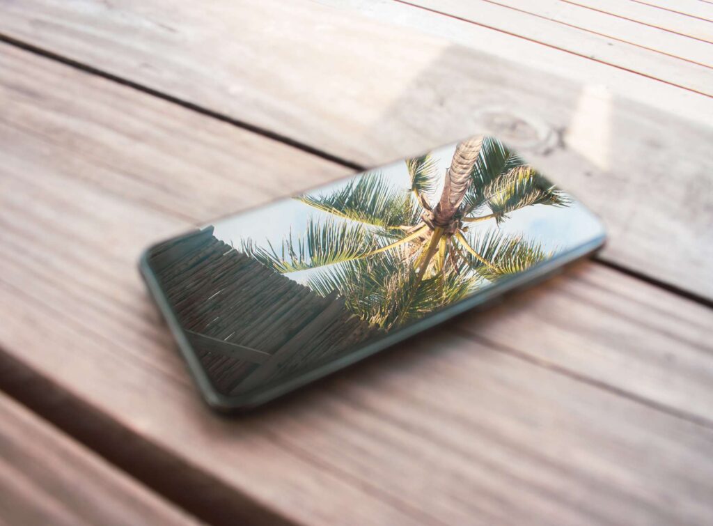 Mobile phone with reflection of palm tree on display. Smartphone on wooden table in tropical resort. Vacations concept. Palm tree reflection at phone screen.