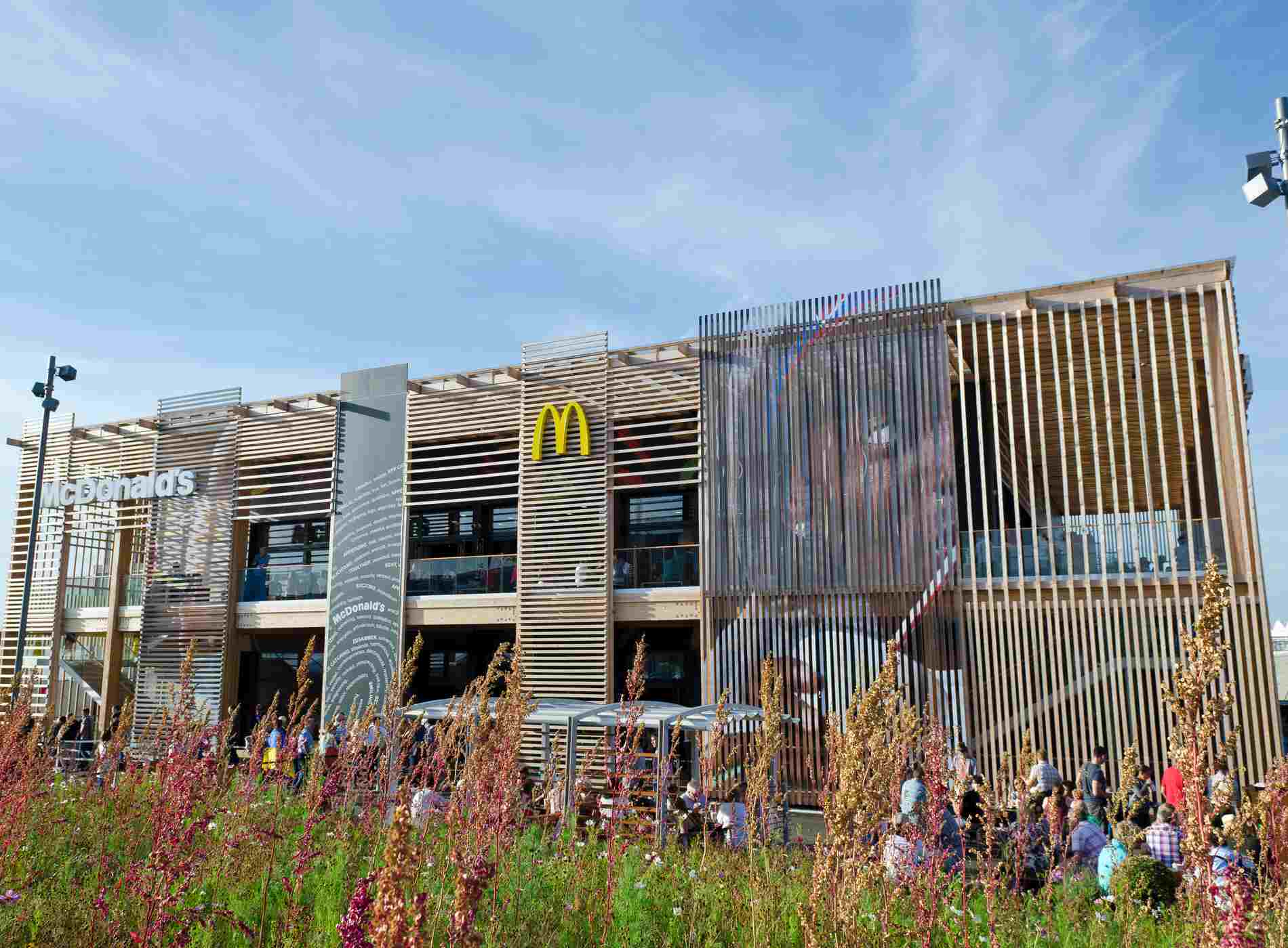 "London, United Kingdom - September 6, 2012: McDonald's flagship restaurant viewed behind a bank of wildflowers. Situated in the London 2012 Olympic Park, it's the largest and busiest McDonald's in the world, the building will be dismantled after the games and has been designed so that three-quarters of the furniture and fittings can be reused in other McDonald's restaurants. People can be seen eating on tables in front of the restaurant and on the balcony of the two storey building."