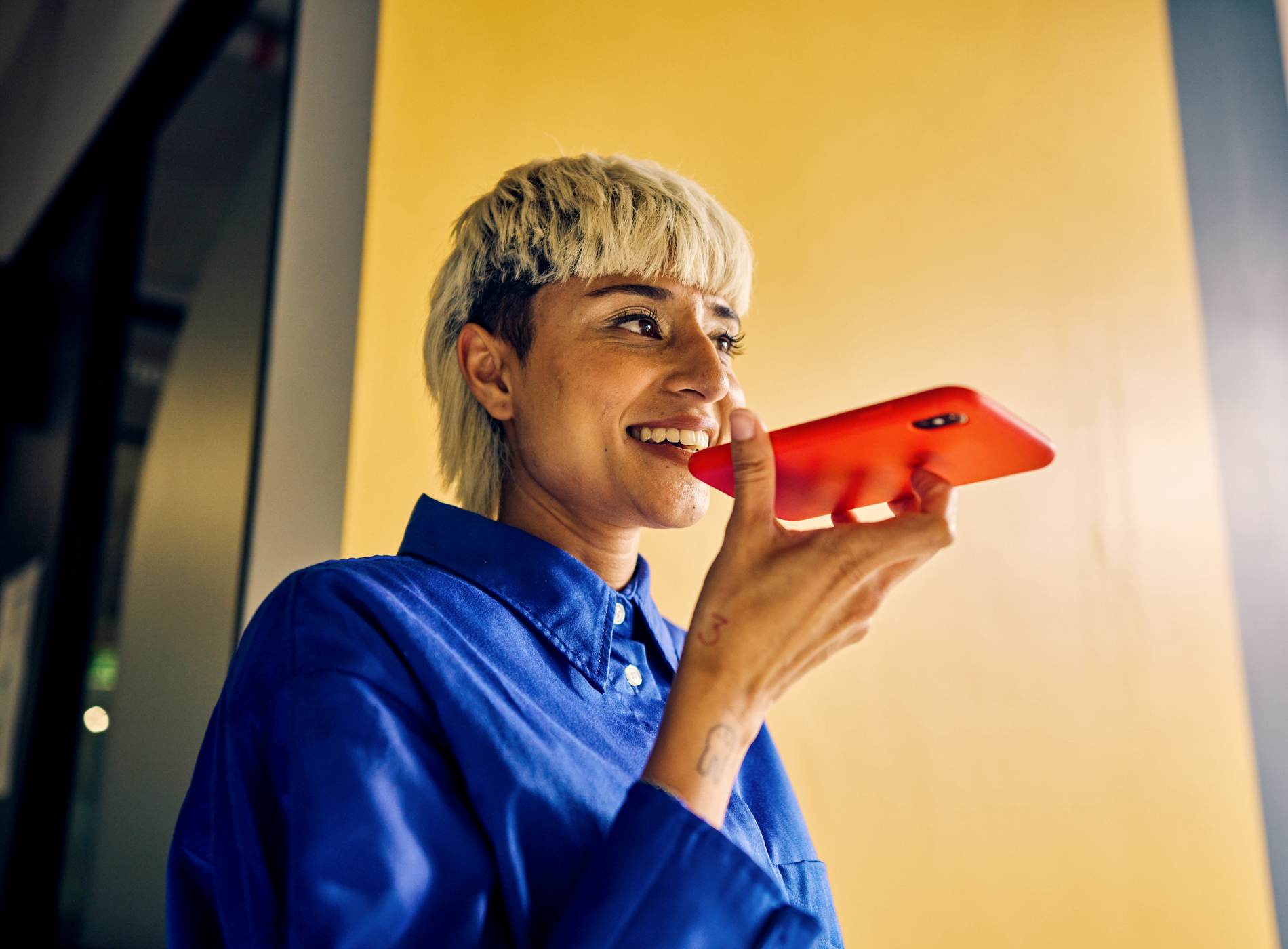 Smiling professional Asian woman using voice assistant on smartphone in a colorful office. Technology and communication in business concept