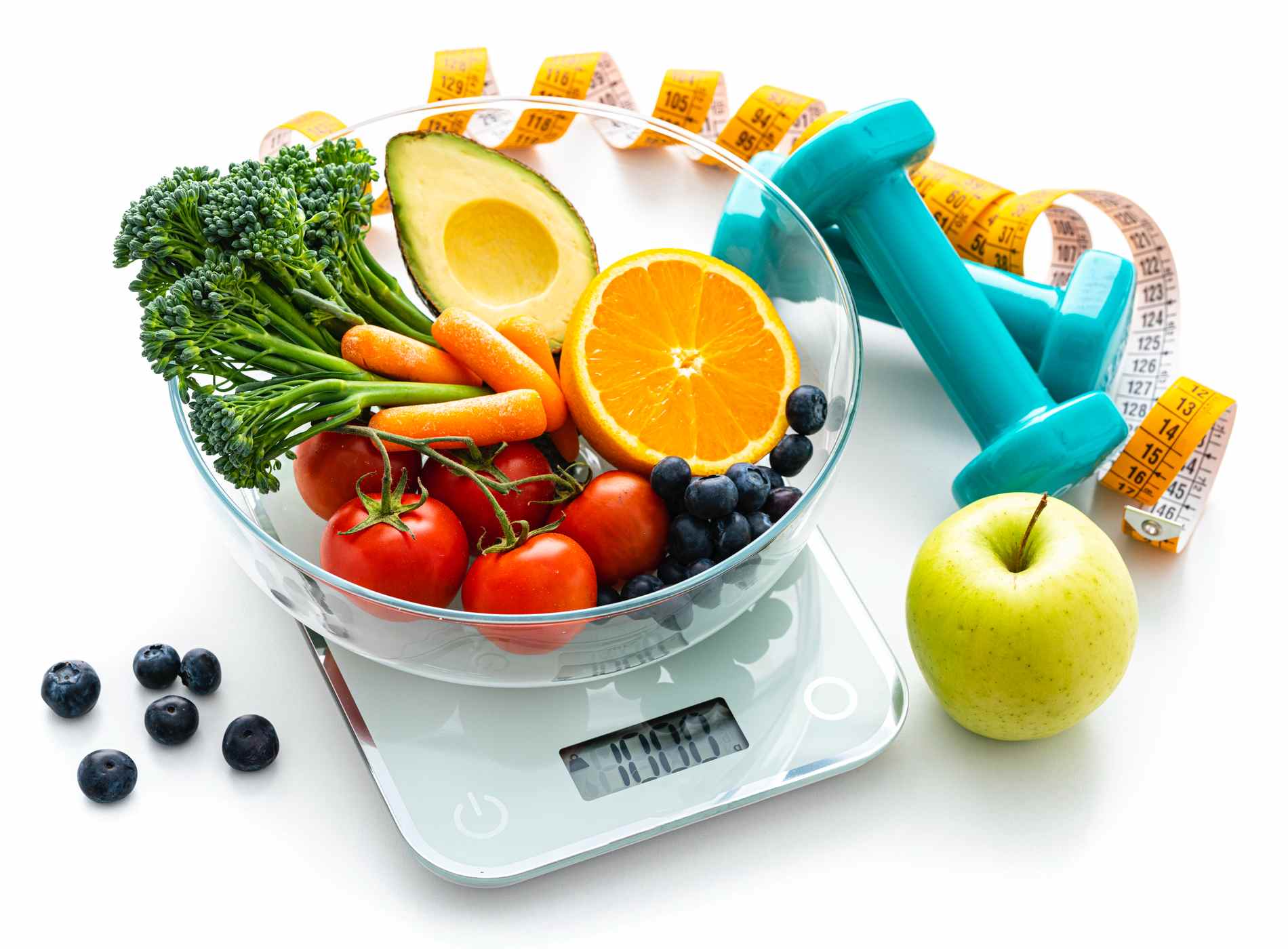 Healthy eating and exercising concept. High angle view of a glass bowl filled with fresh organic fruits and vegetables placed on a kitchen scale isolated on white background. Yellow tape measure and dumbbells complete the composition. The composition includes orange, carrot, broccoil green apple, tomatoes and berry fruits.