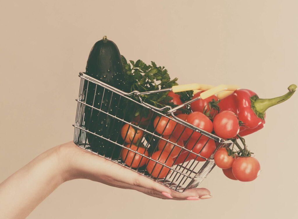 Adult woman do not like to eat vegetables, healthy food, vegetarian products. Female holding small shopping basket with green red vegetables, stop gesture, on grey