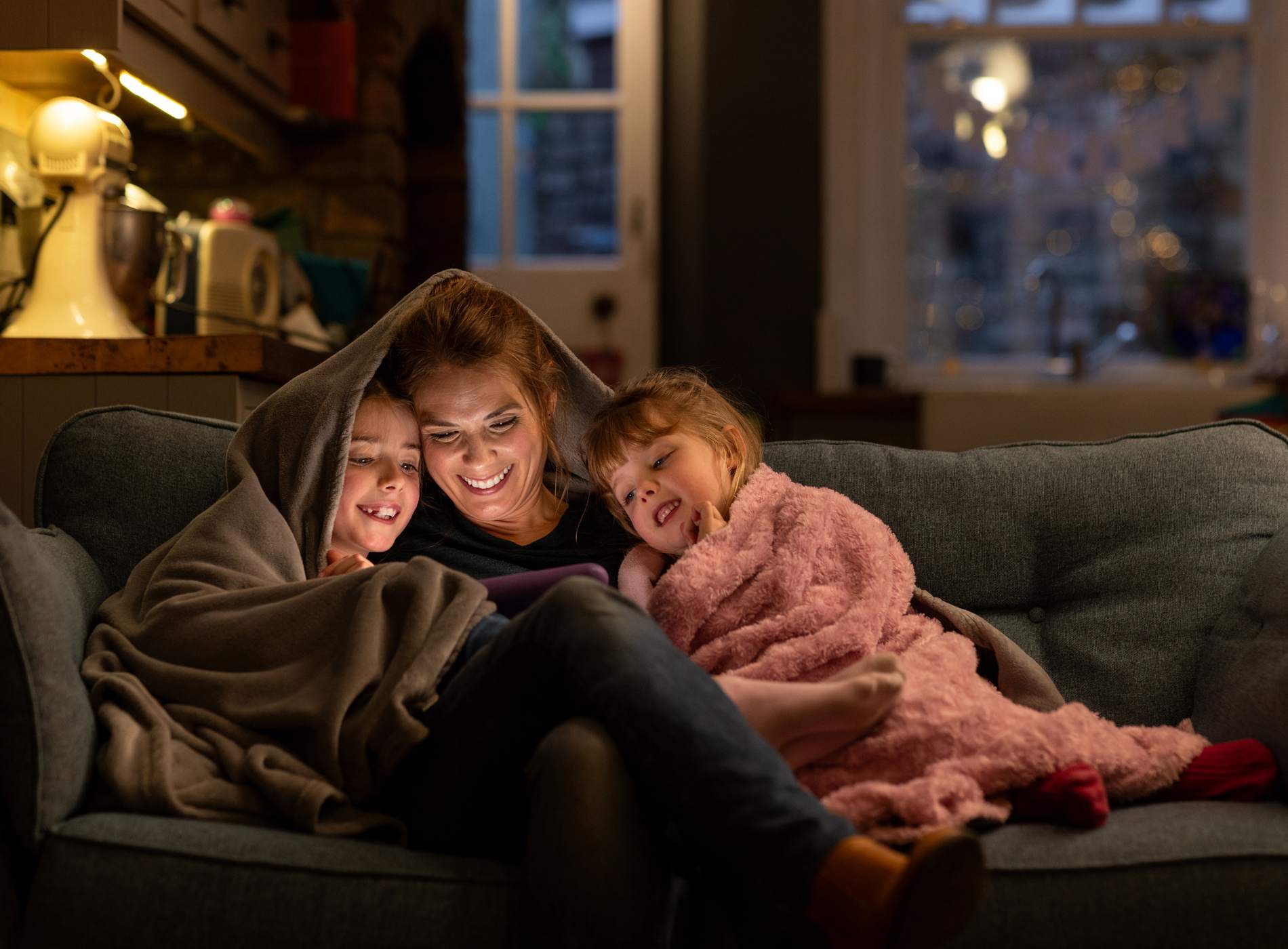 A shot of two young girls with their mother, sitting comfortably with a blanket over their heads on a sofa in the living room of their home in South Shields, North East England. They are all smiling, looking at a digital tablet which the mother is holding. In the background the kitchen is in partial view, and it is dusk outside.

Video similar to this scenario available.