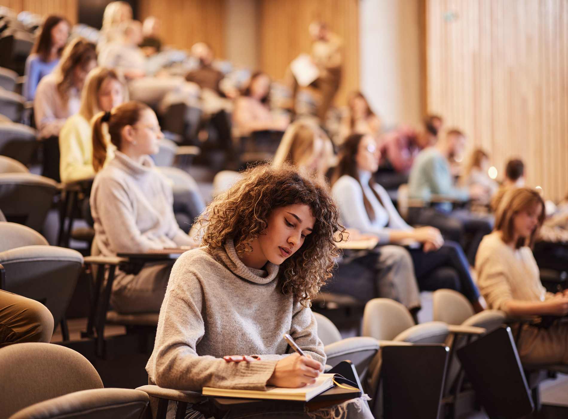 Female college student writing an exam during a class at lecture hall. Her classmate are in the background.